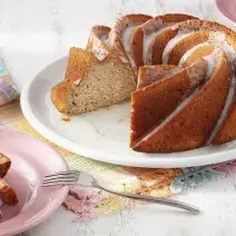 Fotografia em tons de branco e rosa de uma bancada branca com um paninho colorido, sobre ele um prato branco redondo com um bolo. A frente um prato redondo rosa com uma fatia de bolo e um garfo.