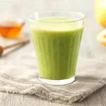 Fotografia em tons de verde em uma bancada de madeira clara com um pano bege, um copo de vidro com o suco de maracujá, couve e mel. Ao fundo, um maracujá cortado ao meio e um potinho com mel.