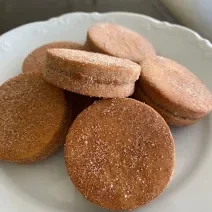 Fotografia em tons de marrom com um prato branco ao centro. Dentro do prato existe algumas unidades de biscoitos de cor caramelo recheados com doce de leite e cobertos com açúcar.