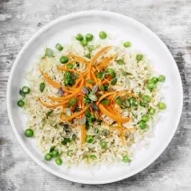 Fotografia vista de cima de um prato branco com arroz, castanha, ervilhas, cenoura e brotos por cima. O prato está sobre uma toalha cinza e branca.