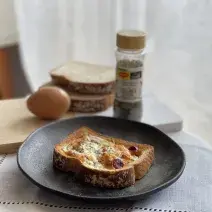 Foto de uma bancada com um prato preto e a receita de Pão com Ovo feito na airfryer. Ao fundo há um pote pequeno de tempero, um ovo e duas fatias de pão sobre uma tábua.