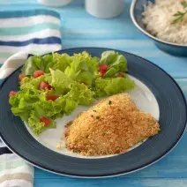 Foto da receita de Filé de Tilápia Copacol com Crosta de Parmesão, vista de cima, em um prato redondo servido com salada, em uma bancada azul com talheresl