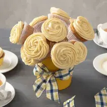 Fotografia em tons de amarelo em uma bancada cinza com um vaso de buquê de cupcake ao centro com um laço xadrez em amarelo e azul. Ao lado, xícaras brancas com chá e pratos brancos com um cupcake em cada um.