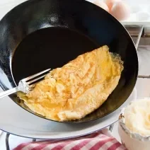 Fotografia de uma omelete dobrada em uma frigideira funda preta que está inclinada para colocar o ovo no prato, e um garfo segundo a omelete. Ao fundo, ovos sobre uma mesa branca. Ao lado do prato que vai receber a omelete, um pano de mesa vermelho.