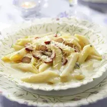 Fotografia tirada de uma prato redondo branco com macarrão e creme branco e pedaços de pinhões