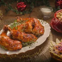 Fotografia em tons de vermelho em uma bancada de madeira escura, um prato oval branco ao centro com as sobrecoxas de frango em cima dele. Ao lado, potinhos com salada e farofa. Ao fundo, decoração e enfeites natalinos.