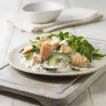 Fotografia de um pepino refrescante e cremoso com salmão defumado e uma salada verde em um prato raso de cerâmica de cor clara. O prato está sobre uma tábua de madeira branca, apoiada em uma mesa de madeira do mesmo tom.