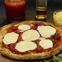 Foto da receita de pizza napolitana servida sobre uma tábua de madeira redonda com folhas manjericão ao lado. Ao fundo, há um bowl de vidro com muçarela, um vidro de azeite, um moedor de sal e um vidro de molho pomodoro