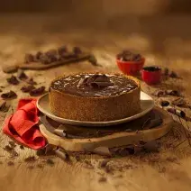 Fotografia em tons de marrom em uma bancada de madeira de cor marrom. Ao centro, uma tábua de madeira contendo o cheesecake e ao redor há pedaços de chocolates espalhados.