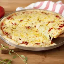 Fotografia em tons de vermelho em uma bancada de madeira clara, uma tábua de madeira, um pano branco com listras vermelhas, uma forma de pizza com a pizza de muçarela em cima dela e uma espátula de madeira tirando um pedaço. Ao lado, tomates para decorar.
