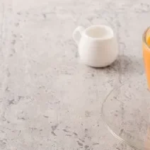 Fotografia em tons claros de uma vitamina laranja com folhas de hortelã por cima em um copo de vidro, apoiado sobre um prato pequeno de vidro. Ao fundo, um pote pequeno branco de vidro, sobre uma mesa em tons de cinza.