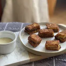 Foto aproximada de um prato com pedaços de Pão com Canela French Toast, ao lado de um potinho pequeno com um pouco de Leite Moça, tudo sobre uma toalha cinza quadriculada