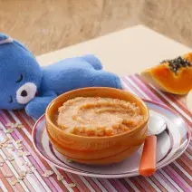 Fotografia em tons de laranja e azul de uma bancada de madeira com um paninho listrado colorido, um potinho laranja com o purêzinho de mamão, atrás um ursinho de pelúcia azul. Ao fundo, meio pedaço de maçã e meio pedaço de mamão.