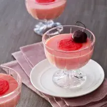 Fotografia em tons de vermelho em uma mesa de madeira com um pano rosa escuro e um prato pequeno branco apoiado nele. Em cima do prato, uma taça de vidro com a mousse de cereja e uma cereja por cima para enfeita. Ao lado e ao fundo, mais duas taças.