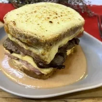 Foto da receita de Francesinha. Oberva0se um prato de cerâmica cinza com o sanduíche por cima, com queijo gratinado e um molho amarronzado.