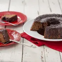 Fotografia em tons de vermelho em uma bancada de madeira clara com um prato branco ao centro com o bolo de chocolate cortado ao meio e um paninho vermelho embaixo. Ao lado, dois pratos vermelhos com uma fatia de bolo em cada um.