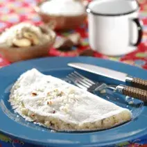 fotografia em tons de azul, branco e vermelho de uma bancada forrada com um pano vermelho e por cima um prato redondo azul com a tapioca e garfo e faca ao lado para servir.