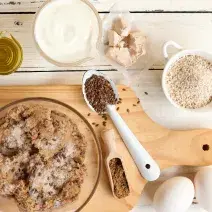 Fotografia em tons de branco e marrom de uma bancada branca com uma tábua de madeira clara, sobre ela um recipiente de vidro com ingredientes. Ao lado dois ovos, e recipientes com azeite, iogurte, fermento e aveia.