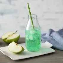 Fotografia em tons de verde em uma bancada de madeira de cor marrom. Ao centro, um prato branco contendo a bebida em cima e ao lado, há uma maçã cortada. Ao fundo, há outra maçã e um pano azul