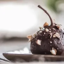 Fotografia de uma pera coberta com chocolate à base de Neston e lascas de nozes sobre um prato de vidro de cor clara.