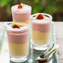 Fotografia em tons de rosa e amarelo em uma bancada de madeira, um recipiente retangular com três taças de vidro com o creme de frutas com o sorvete de creme. Morango e manga para decorar as taças.