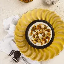 Fotografia em tons de amarelo em uma bancada de madeira branca. No centro, um prato redondo contendo o Bolo Piscina. Ao fundo, um recipiente com poupa de maracujá e um outro com negresco.