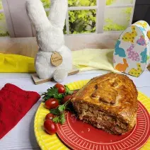 Foto da receita de Ovo de Colher Torta de Frango, servido cortado em um prato vermelho, sobre outro prato amarelo, em uma bancada branca decorada com itens de páscoa, como um coelho branco e uma caixinha em formato de ovo de páscoa