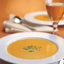 Fotografia em tons de laranja em uma mesa de madeira, com uma taça de vinho branco e um prato branco fundo com a sopa de abóbora, maçã e gengibre dentro dele.