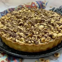 Foto da receita de Torta Nocciolíssima. Observa-se uma torta inteira decorada com avelãs e creme de chocolate sobre um prato de cerâmica redondo preto.