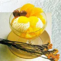 Fotografia em tons de amarelo em uma bancada de madeira, um pano bege, um prato redondo raso bege com uma taça de vidro com o sorvete de creme e pêssegos em calda dentro dela. Ao lado, flores amarelas e laranjas.