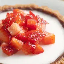 fotografia em tons de branco e vermelho tirada de um prato redondo branco que contém uma mini-torta com creme branco e pedaços de morangos com um garfo ao lado para servir.