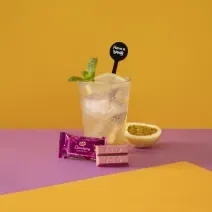 Foto da receita de Soda Italiana Passion Fruit and Lemon. Observa-se um fundo amarelo e roxo com um copo alto no centro, cheio de gelo, com uma rodela de limão siciliano. Meio maracujá decora a foto à direita, assim como folhas de hortelã no copo.