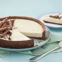 Fotografia em tons de azul em uma mesa de madeira azul com um prato raso azul claro e a torta de limão e chocolate. Ao lado, um prato pequeno com uma fatia da torta e uma espátula de bolo.