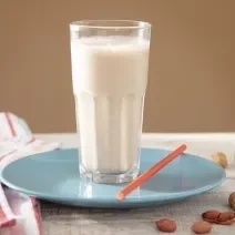 Foto em tons claros da receita de vitamina proteica com pasta de amendoim servida em um copo alto sobre um prato de porcelana azul claro com um canudo vermelho ao lado. Ao fundo há um pano colorido além de amendoins espalhados