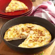 Fotografia em tons de vermelho em uma bancada de madeira, uma tábua de madeira, um prato redondo fundo com a omelete dentro dele. Ao lado, pratos pequenos vermelhos com uma fatia da omelete.