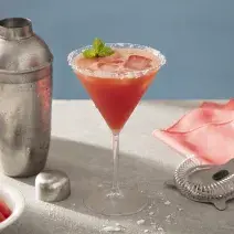 Foto em tons de vermelho da receita de margarita de melancia servida em uma taça de vidro sobre uma mesa de mármore onde estão uma coqueteleira cromada, além de um pano vermelho, uma mola para mistura, um copo com gelo e pedaços de melancia em cubos.