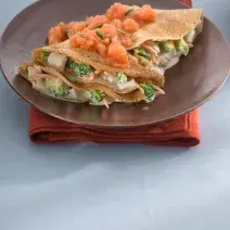 Fotografia de um prato marrom com 2 panquecas feitas em forma de crepe e recheadas com legumes e cobertas com molho de tomate, sobre guardanapo vermelho e toalha azul clara.