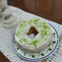 Foto em tons de branco da receita de bolo de nescafé com cobertura de limão servida em uma porção grande sobre um prato decorado em cima de uma mesa de madeira com um pano bordado