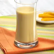 Fotografia em tons de laranja em uma bancada de madeira com uma toalha colorida listrada, um pano laranja e um copo de vidro alto com a vitamina de banana com cupuaçu. Ao fundo, um pratinho branco raso com fatias de manga.