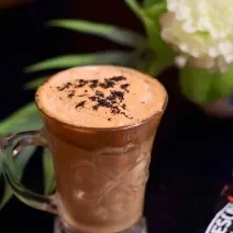 Fotografia de uma xícara de vidro com uma bebida com chocolate, Nescafé e leite. Ao lado, uma embalagem de Nescafé, e ao fundo, decoração de plantas e flores, sobre uma mesa preta.