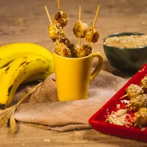 Foto da receita de Banana e Neston no espeto, com alguns espetos dentro de uma caneca amarela e outros em um prato, tudo em uma bancada de madeira decorada com bananas, trigo e um caneca com Neston 3 Cereais dentro.