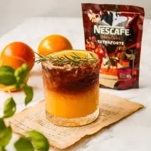 Fotografia em tons de laranja em uma bancada de mármore com um copo de vidro ao centro e o suco de laranja com café dentro dele, decorado com um ramo de alecrim. Ao fundo, laranja e embalagem de café.