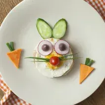 fotografia em tons de marrom, branco, laranja e verde tirada de uma bancada marrom vista de cima. Ao centro um prato redondo branco com um sanduiche em formato de coelho.