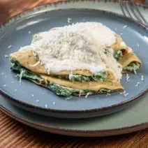 Fotografia de dois crepes de queijo e espinafre dobrados com molho branco por cima e queijo ralado. Os crepes estão em um prato azul raso, que está sore outro prato, um pouco maior, e ao lado, um garfo apoiado.