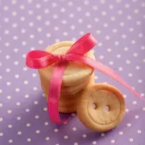 fotografia em tons de roxo, bege e rosa de uma bancada roxa com bolinhas brancas, contém biscoitinhos em formatos de botão com um laço rosa por cima.