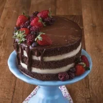 Imagem da receita de Naked Cake de Cacau, decorado em camadas marrons e brancas, com chocolate escorrendo, decorado com frutas vermelhas. O bolo está sobre um prato azul em uma bancada de madeira.