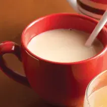 Fotografia em tons de vermelho, branco e marrom, com xícara vermelha contendo uma bebida em tom de branco, no entorno outras duas xícaras, tudo sobre bancada em tons de marrom.