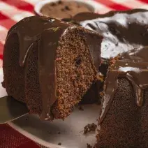 Foto bem aproximada de um bolo de chocolate com uma fatia sendo retirada. O bolo está num prato branco e coberto com Brigadeiro e o prato está sobre uma toalha xadrez vermelho e branco.