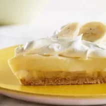 Fotografia de uma fatia de torta de mousse de banana, merengue e rodelas de banana por cima sobre um prato amarelo.