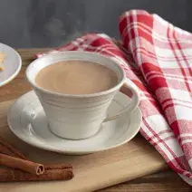 Fotografia em tons de vermelho e marrom de uma bancada, ao centro uma xícara com chocolate quente saindo fumaça e um pires abaixo. Ao lado pedaços de canela, biscoitinhos e uma pano xadrez vermelho e branco.